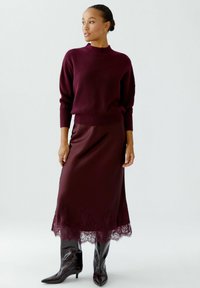 Pull en tricot bordeaux à col montant, associé à une jupe satinée bordeaux avec des détails en dentelle sur l'ourlet. Fini avec des bottes noires brillantes.