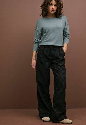 Maglione azzurro chiaro a maniche lunghe con polsini a costine, abbinato a pantaloni larghi a righe nere e scarpe beige slip-on, su uno sfondo marrone.