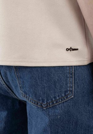 Nærbilde av beige skjorteskjørt med liten svart brodert logo over baklommen på blå jeans.
