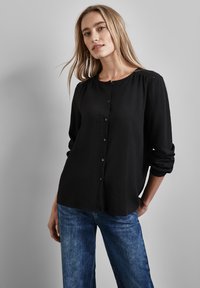 Blusa nera a maniche lunghe con chiusura a bottoni, tessuto testurizzato, collo rotondo e spalle arricciate, abbinata a jeans larghi blu.