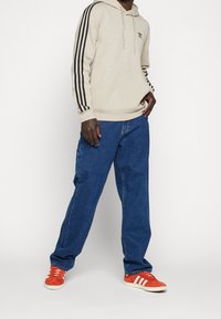 Person som bär en beige Adidas huvtröja med svarta ränder, blå jeans och röda Adidas sneakers med vita ränder, stående mot en vit bakgrund.