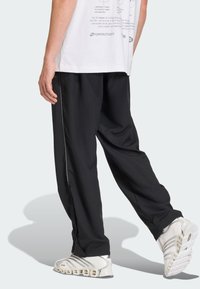 Mann iført svarte, løstsittende bukser, hvit t-skjorte med trykk og hvite Adidas sportssko med hul såle, stående på lys bakgrunn.