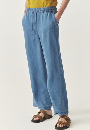 Helle blaue, weit geschnittene Hose aus Denim-Stoff mit einem elastischen Bund und Kordelzug. Verfügt über Seitentaschen. Getragen mit braunen Sandalen.