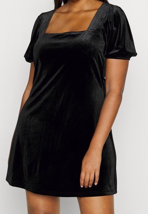 Kobieta ubrana w krótką czarną aksamitną sukienkę z bufkowatymi krótkimi rękawami i kwadratowym dekoltem, stojąca na białym tle.