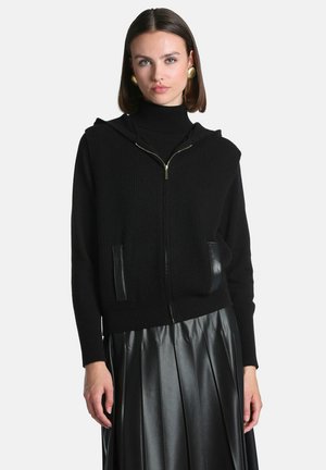 Femme portant un sweat à capuche noir zippé avec des détails en cuir aux poches et une jupe plissée en cuir noir sur un fond uni.
