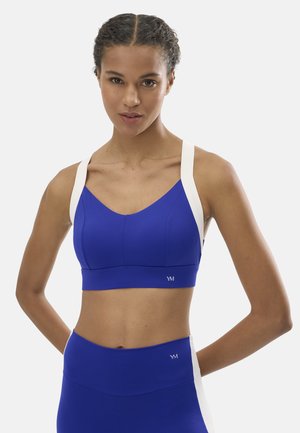 Mujer vestida con un sujetador deportivo azul y leggings de talle alto con tiras blancas, de pie con las manos detrás de la espalda contra un fondo liso.