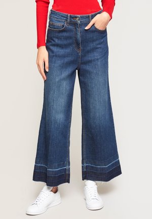 Persona que lleva puestos vaqueros de pierna ancha y corte cropped en azul oscuro con dobladillos en contraste, una camiseta roja de manga larga y zapatillas blancas, de pie frente a un fondo liso.