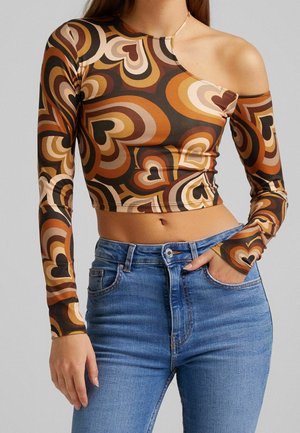 Kvinde iført en farverig retro crop top med hjerteprint og et skulder samt højtaljede blå jeans med den ene hånd i lommen.