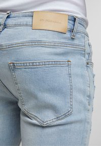 Jean en denim bleu clair avec une poche arrière et une étiquette en cuir beige portant la mention "2Y PREMIUM", porté avec un haut à côtes blanc.