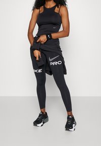 Donna che indossa leggings Nike Pro neri, una canotta nera, un braccialetto Nike nero, sneakers nere e una giacca nera legata in vita.