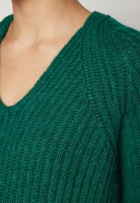 Gros plan d'une personne portant un pull vert à col en V en maille avec une texture côtelée épaisse, montrant la zone du cou et des épaules.