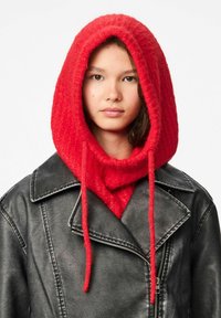Capucha roja de punto con cordones largos, llevada sobre una chaqueta de cuero negra con solapas anchas y cierre con cremallera. Tejido texturizado.