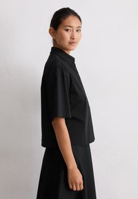 Camicia nera a maniche corte con colletto a punta e design corto, realizzata in tessuto liscio, con chiusura a bottoni e cuciture minime.