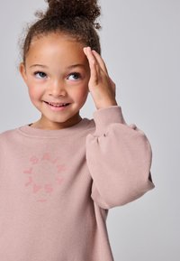 Hymyilevä nuori tyttö, jolla on kiharat hiukset nutturalla, pukeutuneena vaaleanpunaiseen röyhelöhihalliseen collegepaitaan, koskettaa päänsä sivua ja katsoo ylöspäin.