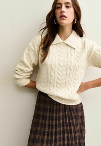 Dicker cremefarbener Zopfmuster-Pullover mit Kragen, kombiniert mit einem braunen, gefältelten Karottenrock mit dunkelblauen und burgunderfarbenen Akzenten.