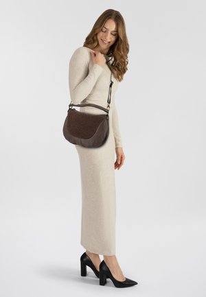 Bruine leren tas met getextureerde accenten, gebogen vorm en korte handgreep. Model draagt een lange beige jurk en zwarte hakken, gepositioneerd op de zijkant.