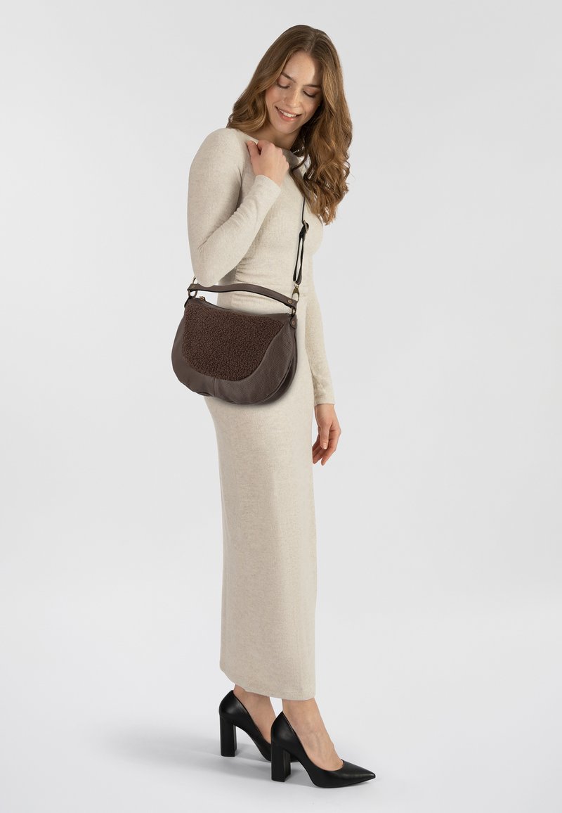 Bruine leren tas met getextureerde accenten, gebogen vorm en korte handgreep. Model draagt een lange beige jurk en zwarte hakken, gepositioneerd op de zijkant.