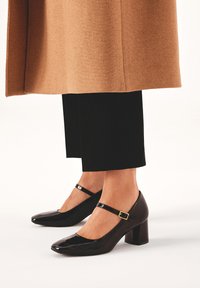 Črne lakirane Mary Jane pete s kvadratnim nosom in blok peto, ki imajo anklet s zlatim zaponko.