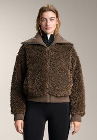 Bruine shearling jas met een hoge kraag, ritssluiting en geribbelde manchetten. De textuur is pluizig en het heeft een relaxte pasvorm.