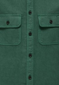 Žalio audinio marškiniai priekyje su juodomis sagomis ir dviem užsegamais krūtinės kišenėmis.