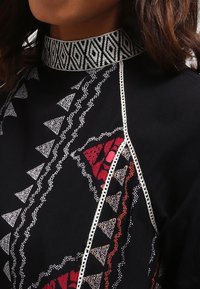 Närbild av en person som bär ett svart plagg med intrikata vita och röda mönster, med en dekorerad mönstrad krage.