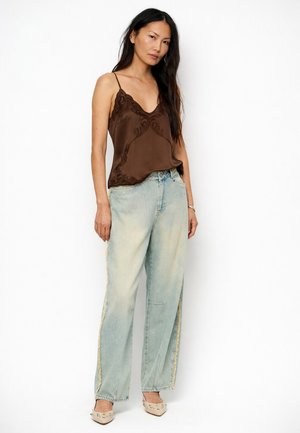 Vrouw die een bruin hemdje met kant, lichtblauwe wijde jeans met versleten zijkstrepen en beige puntige platte schoenen met studs draagt, staand tegen een witte achtergrond.