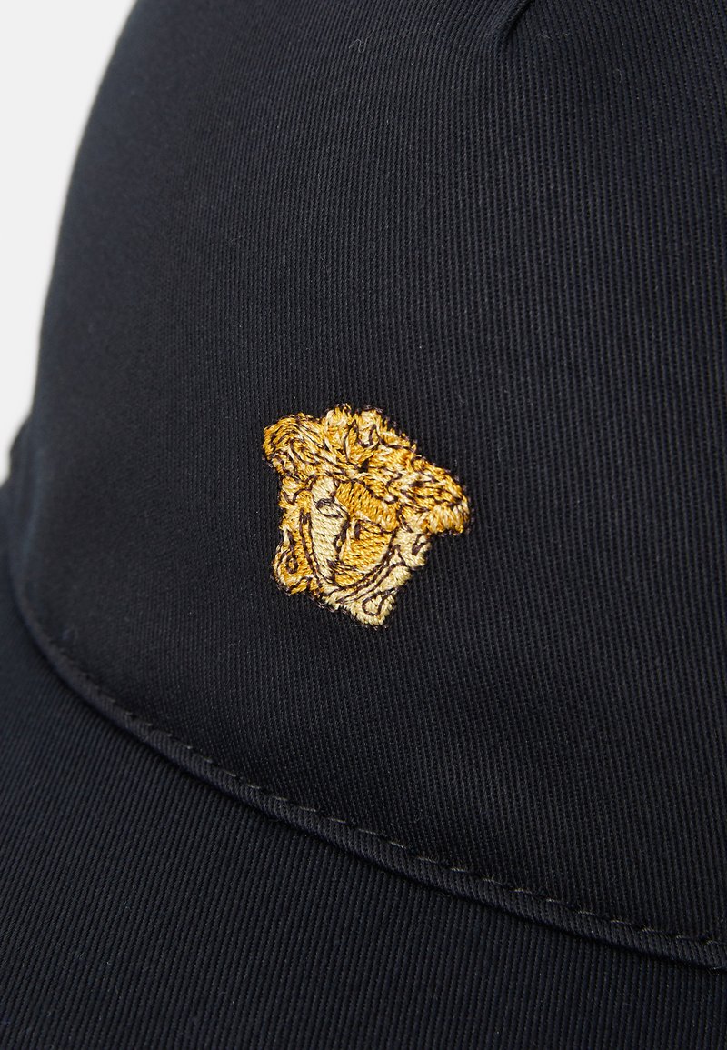 Zwarte pet gemaakt van een gestructureerde stof, met een gouden geborduurd embleem van een Medusa-kop aan de voorkant. Naaddetails zichtbaar op de rand.