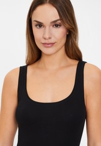 Juoda be rankovių bluzonė, pagaminta iš minkštos medžiagos, su kvadrato formos iškirpte ir plačiomis petnešėlėmis, lygiu paviršiumi ir minimaliais siuvimo detalėmis.