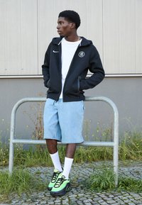 Sort lynlås Hoodie med logo, hvid t-shirt, lyseblå denim shorts, hvide sokker og sort/grønne sneakers. Står ved et metalgelænder.