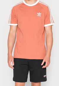 Krátký rukáv, korálové bavlněné tričko s bílým lemem a třemi bílými pruhy na ramenou; na hrudi se nachází malé bílé logo Adidas. Černé šortky pod tím.