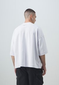 Hvit oversized t-skjorte med korte ermer laget av myk stoff, med en liten grå logo på baksiden, har en avslappet passform og bred halsåpning.