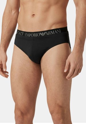 Uomo che indossa slip neri Emporio Armani con fascia in vita con logo, mostrato dalla vita a metà coscia su sfondo neutro.
