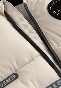 Beige gepolsterte Weste mit schwarzem Reißverschluss und Akzenten. Enthält ein sichtbares Logopatch und das Wort "BAKER" in Hellgrau auf dem Stoff.