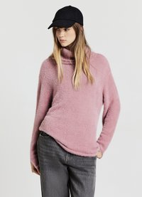 Maglione rosa soffice con collo ampio e maniche lunghe, abbinato a jeans grigi e un cappello nero. La texture appare morbida e confortevole.