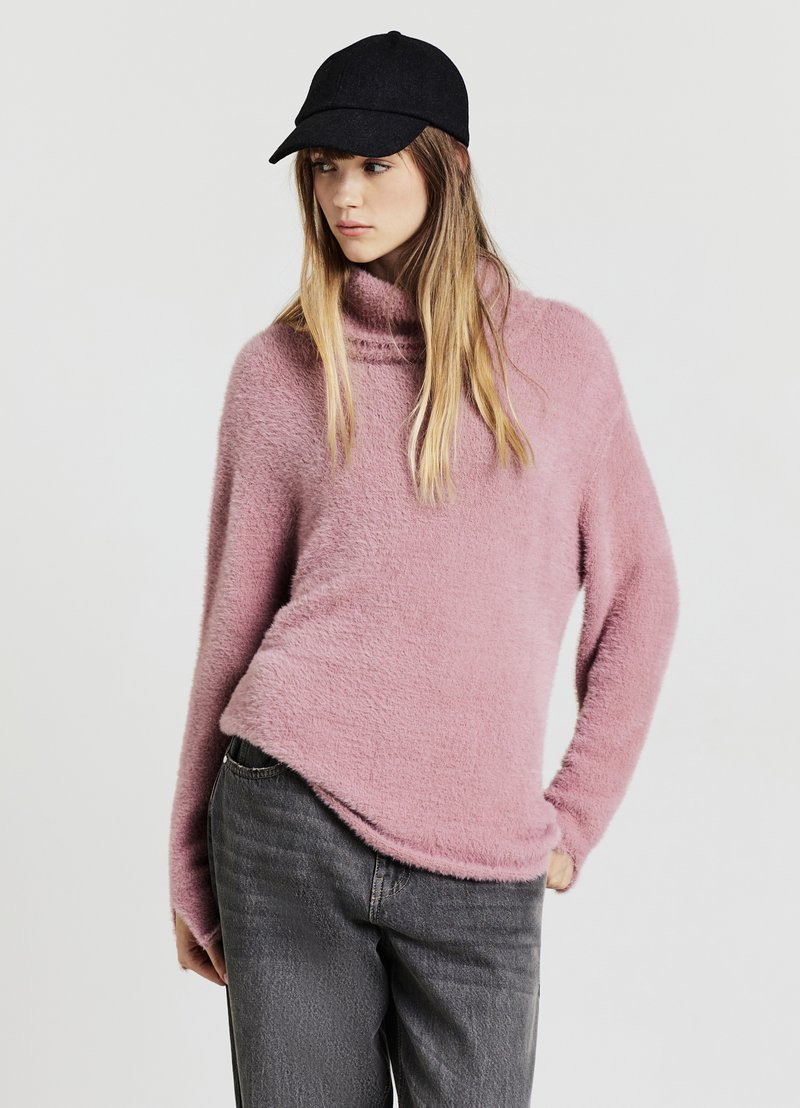 Maglione rosa soffice con collo ampio e maniche lunghe, abbinato a jeans grigi e un cappello nero. La texture appare morbida e confortevole.