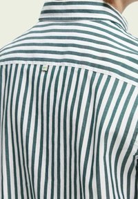 Koszulka w paski z zielonymi i białymi pionowymi paskami, kołnierzykiem i małą metką z tyłu. Tekstura materiału wydaje się miękka i gładka.