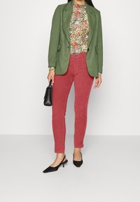 Blazer a quadri verdi sopra una maglietta floreale, abbinato a pantaloni rossi e décolleté nere a punta. Le caratteristiche includono una vestibilità sartoriale e tessuti texturizzati.