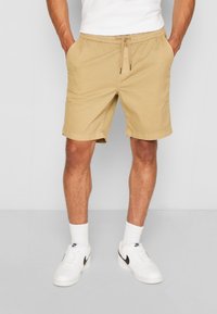 Homme portant un short beige à cordon de serrage, des baskets Nike blanches avec des swooshes noirs, et des chaussettes blanches montantes, se tenant debout les mains dans les poches.