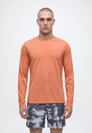 Un homme porte un haut de sport orange à manches longues et un short à motifs noirs et blancs, sur un fond clair uni.