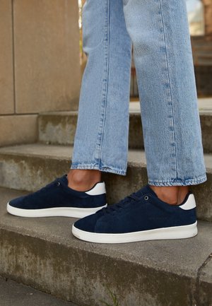 Person iført lyseblå jeans og marineblå ruskindssneakers med hvide såler står på betontrapper udendørs.