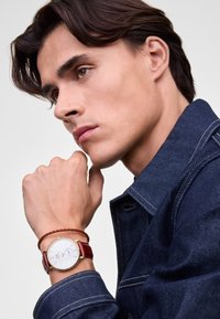 Reloj de cuero marrón con un dial blanco y acentos dorados en una muñeca, acompañado de una pulsera trenzada marrón. El modelo lleva una chaqueta de mezclilla azul.