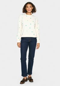 Sudadera de cuello redondo en marfil con patrones de corazones verdes, combinada con jeans azul oscuro y mocasines marrones. Tejido suave y ajuste relajado.