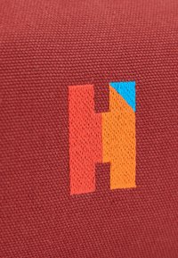 Tissu rouge avec une grande lettre H brodée en rouge et orange, avec un petit triangle bleu dans le coin supérieur droit du H.