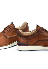 Zapatos de cuero marrón con un cuello de tela texturizada, punta redonda, suela blanca en contraste y acento rojo alrededor de los ojetes.
