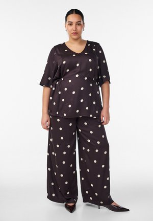 Donna che indossa un completo in raso marrone scuro con pois bianchi, composto da una maglia ampia e pantaloni a gamba larga, in piedi su sfondo bianco.