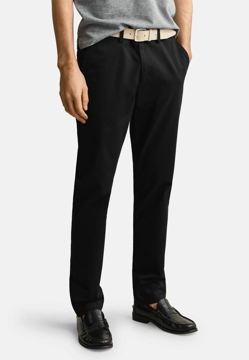 Mann trägt schwarze formelle Hose, schwarze Loafer, graues hineingestecktes Hemd und hellbeigen Gürtel, steht mit den Händen in den Taschen.