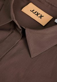 Camicia marrone con colletto realizzata in tessuto liscio con un'etichetta che mostra "JJXX." Presenta un design a colletto classico e una texture uniforme.
