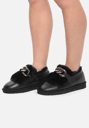 Piernas con zapatos negros tipo slip-on con parte superior peluda y grandes detalles de cadena plateada, sobre un fondo gris claro.