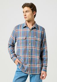 Camisa de franela a cuadros en tonos azul, verde y rojo con cuello, cierre de botones y dos bolsillos en el pecho. Textura suave y mangas largas.