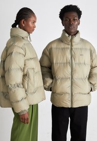 Chaquetas tipo puff en verde oliva claro, con diseño acolchado, cuellos altos y bolsillos laterales. Modelo a la izquierda con un corte corto.